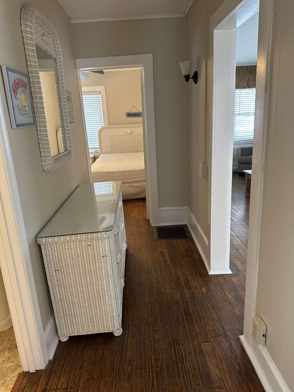 Narrow hallway with dark hardwood floors, white wicker dresser topped with glass, and a wall mirror.
