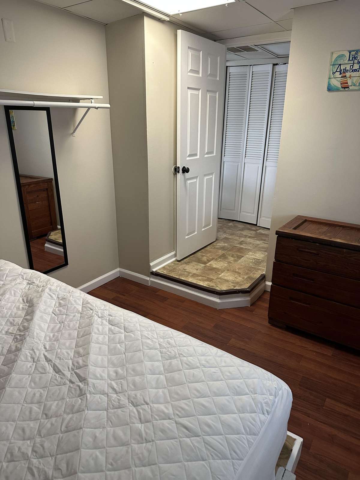 White quilted bedspread covers mattress beside a dark wood dresser and full-length mirror on light gray walls.