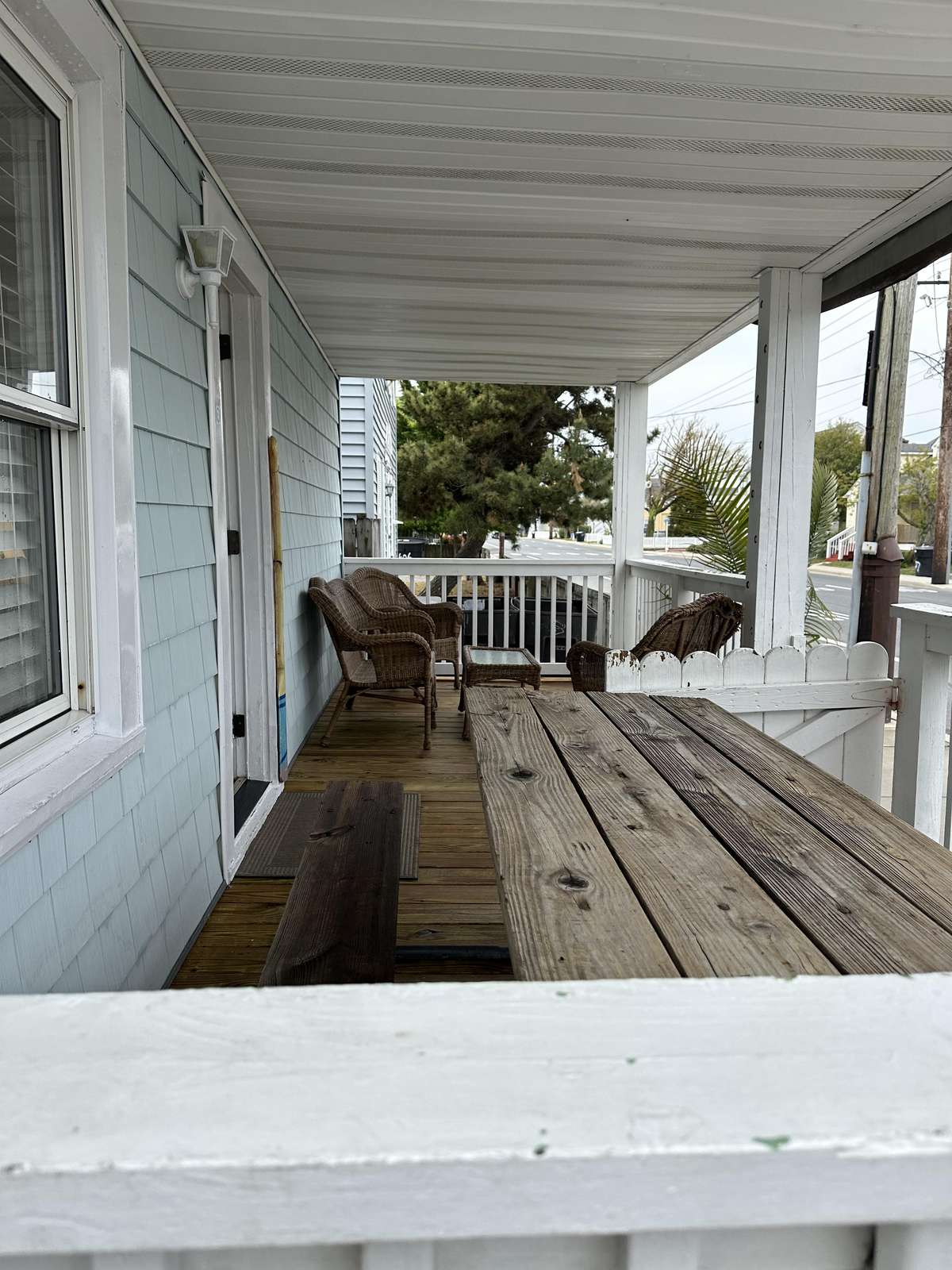 Covered front porch with weathered wood picnic table, wicker chairs, and light blue exterior siding.