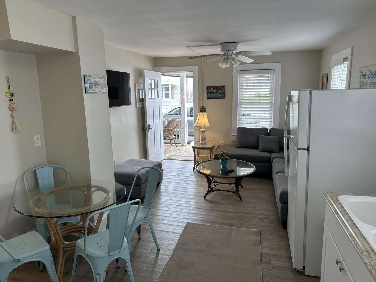 Glass-top dining table with four metal blue chairs opens to a gray sectional and wicker coffee table near the front porch.