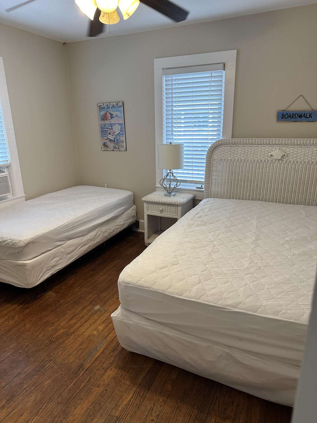 Bedroom with white wicker headboard, two beds with quilted covers, hardwood floors, and coastal wall decor.
