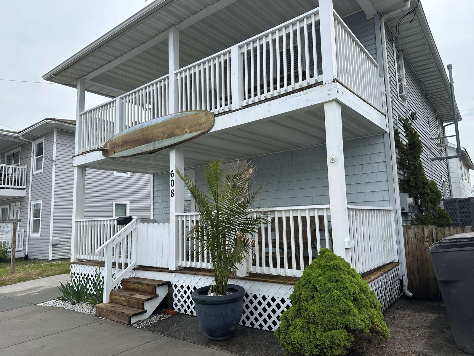 Two-story cottage exterior with white railings, wood steps, blue siding, and a mounted vintage surfboard.