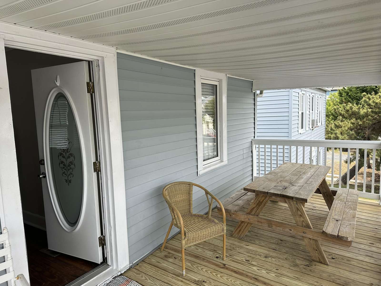 Covered porch with natural wood picnic table, wicker chair, and pale blue vinyl siding under white ceiling.
