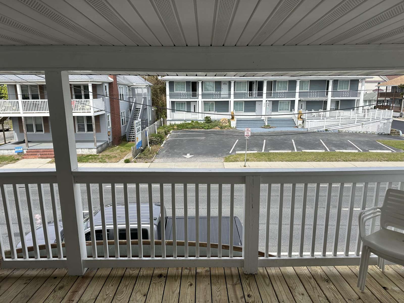 Front porch with natural wood flooring, white railings, and stacked white plastic chairs overlooking the street and neighboring buildings.