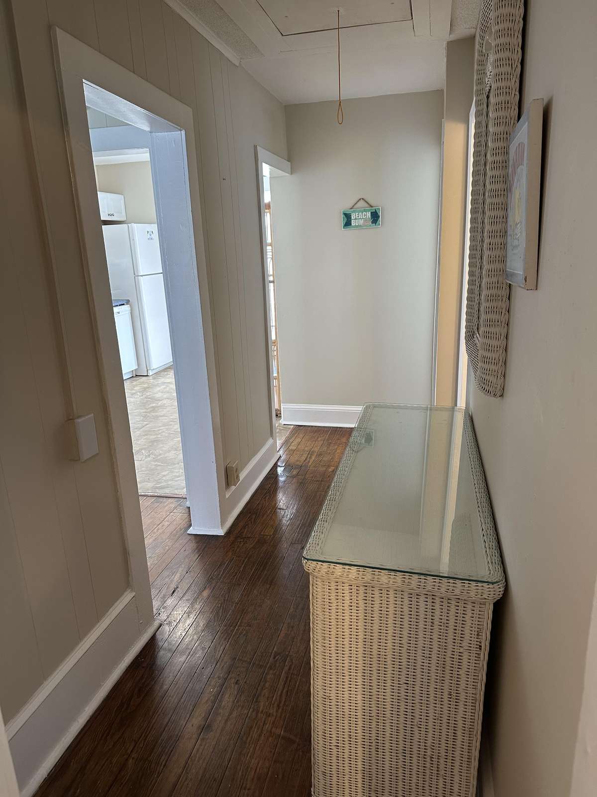 Narrow hallway features dark hardwood floors, light gray walls, white trim, and a glass-topped wicker console.