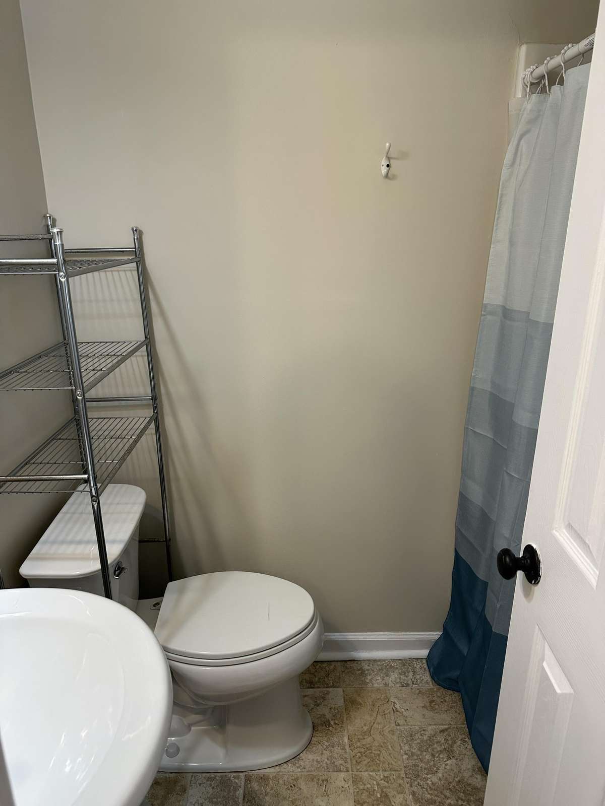Bathroom corner with white toilet, pedestal sink, metal shelving unit, and blue-gray striped shower curtain.