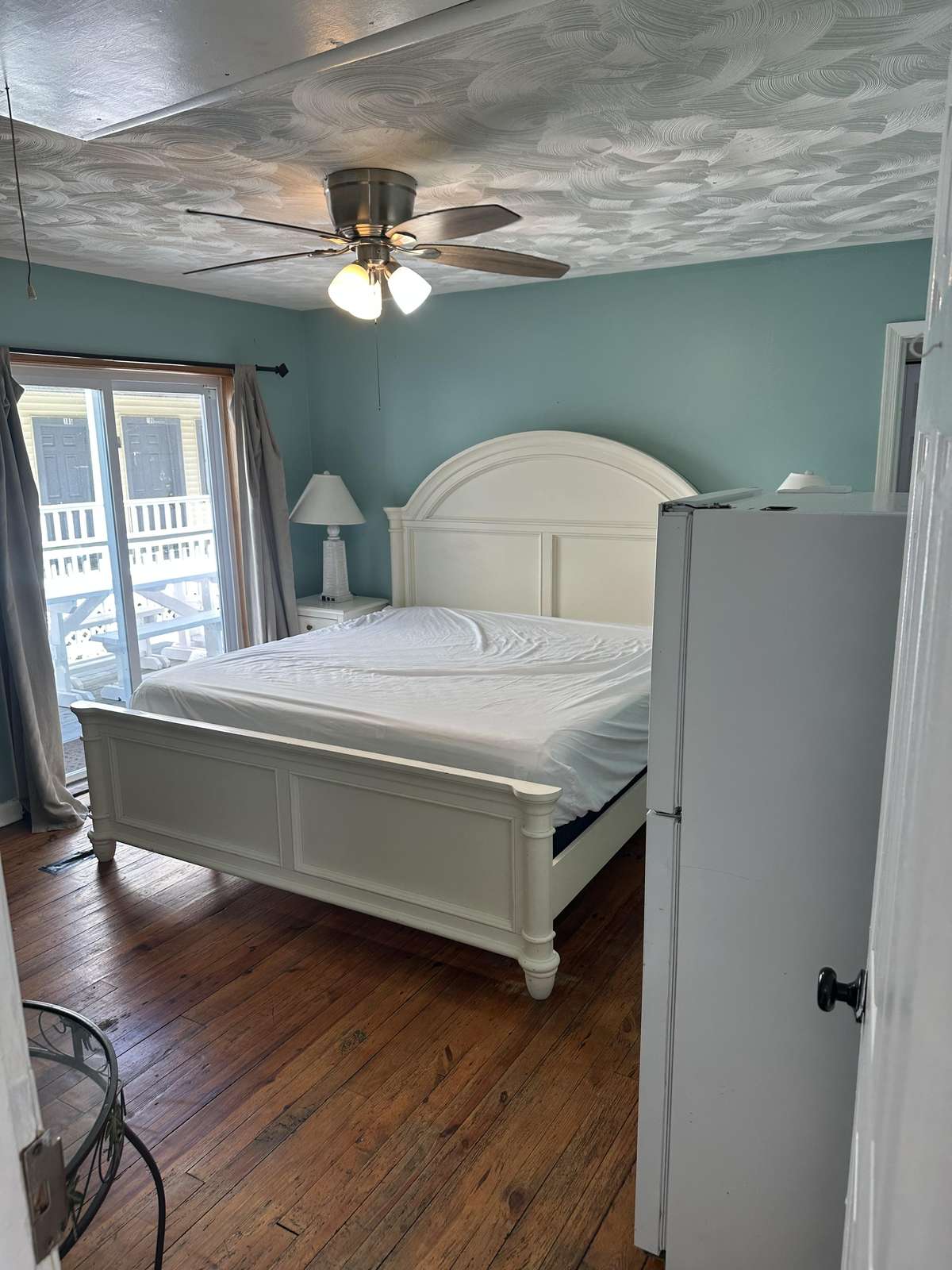 King bed with white paneled headboard sits on polished hardwood floors beneath a ceiling fan and blue walls.