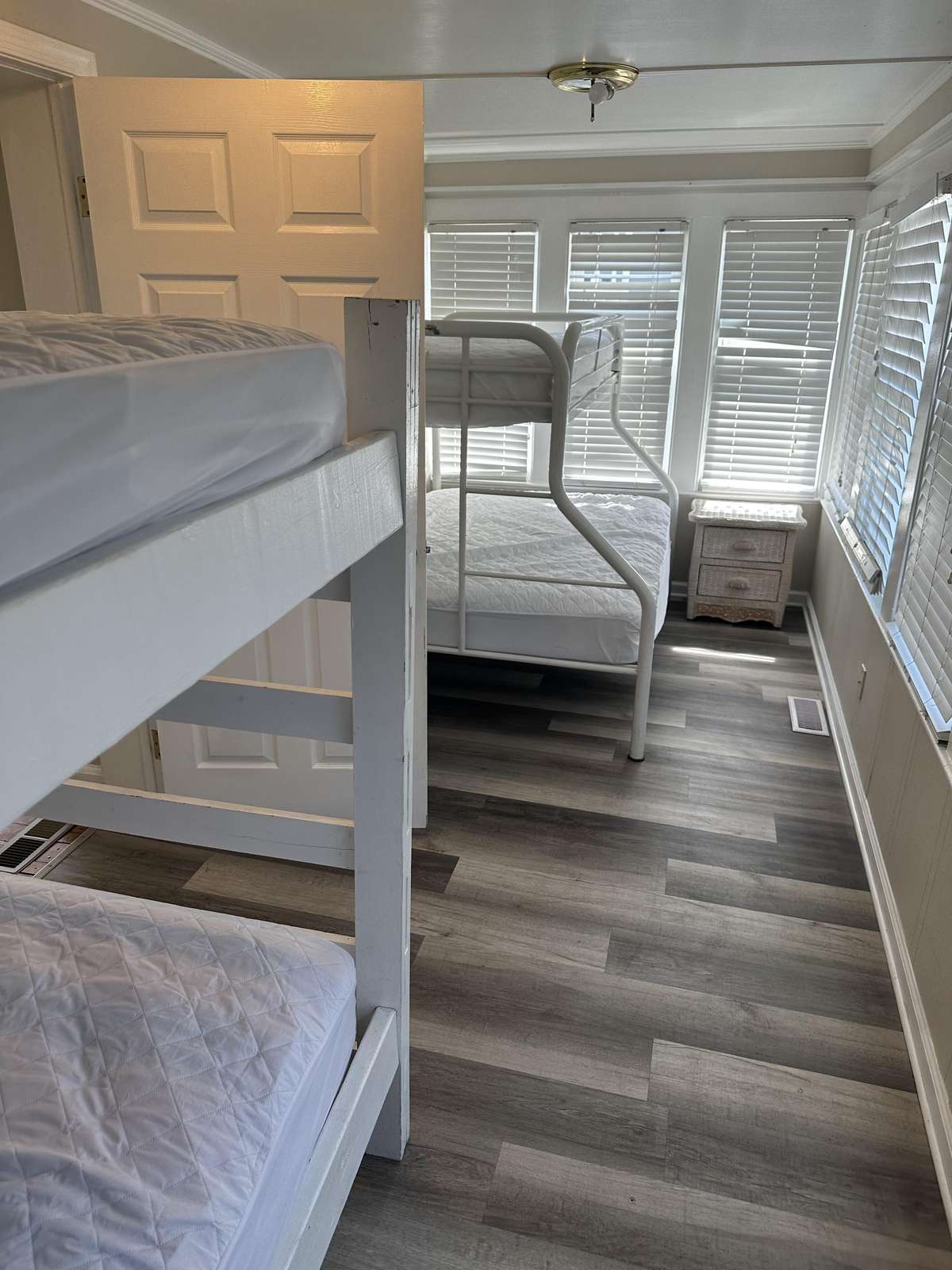 Two white bunk beds and a wicker nightstand fill the bedroom with gray wood flooring and multiple blinds-covered windows.