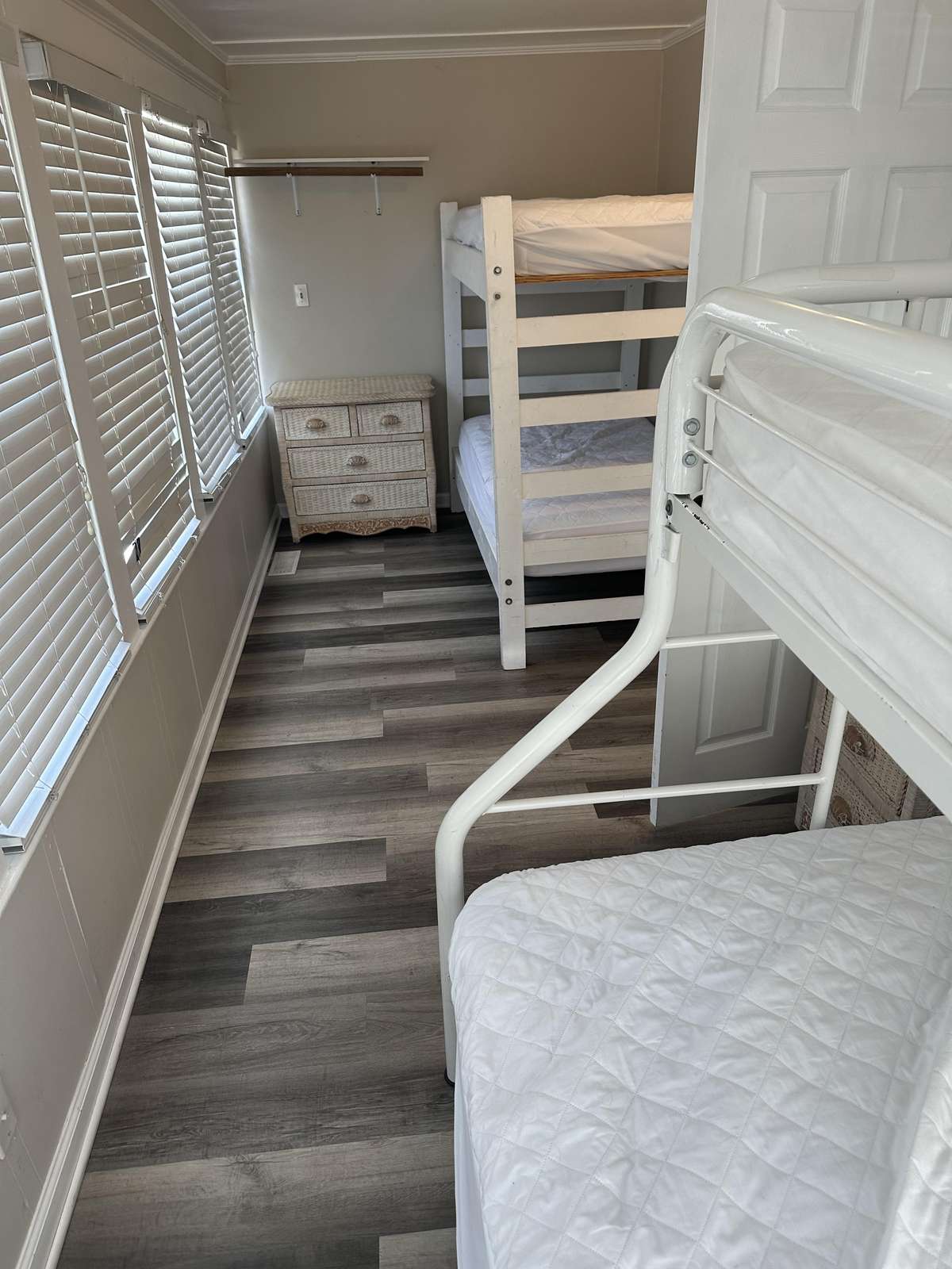 Two white bunk beds with quilts face a wicker chest under a wall shelf beside shuttered windows.
