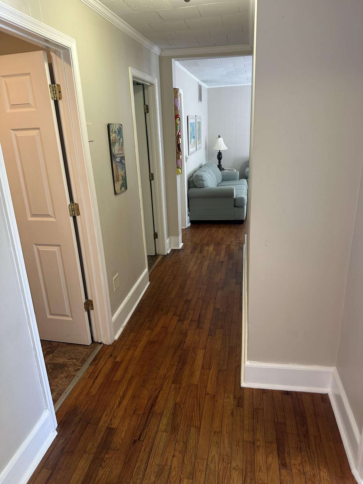 Hardwood floor hallway leads past white-trimmed doorways toward a living area with pale blue couches and a black lamp.