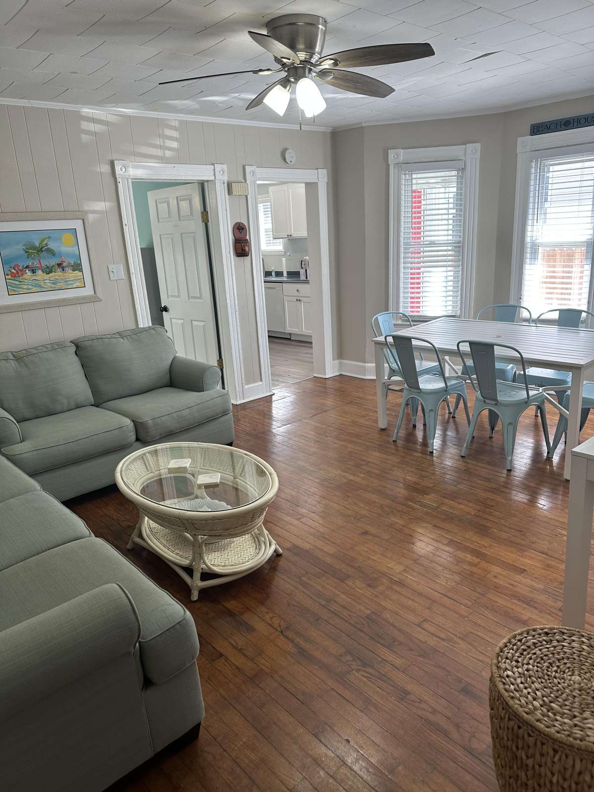Living room with sage green sofas, woven glass-top coffee table, and adjacent dining area with metal chairs.