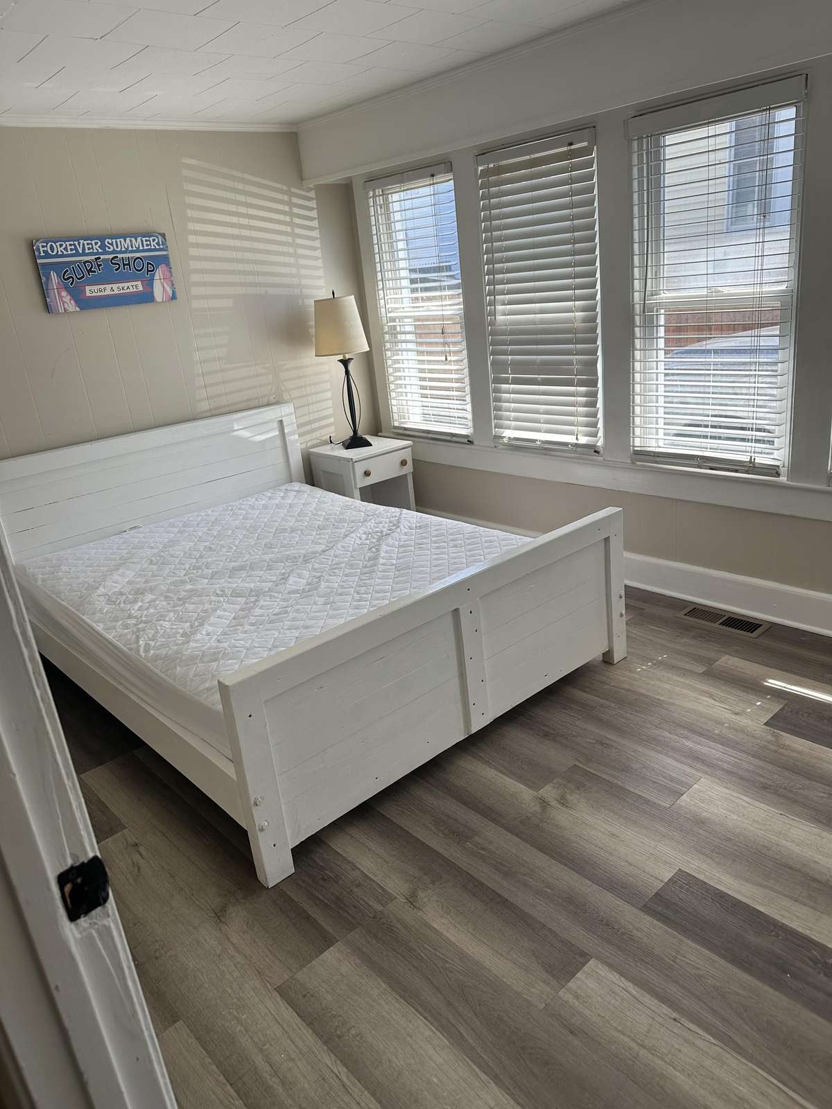 A white wooden bedframe anchors the bedroom with light wood flooring and three tall windows with blinds.