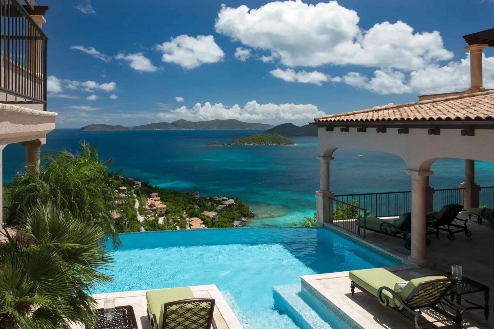 Infinity pool edged with pale stone and green cushioned loungers overlooks tropical hillside and distant islands. - property