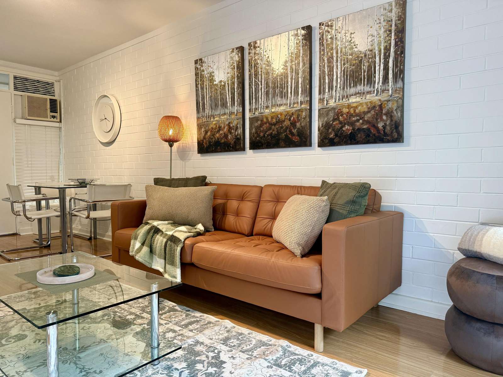 Lounge area with tan leather sofa, knitted pillows, glass coffee table, and forest-themed triptych on white brick wall. - property