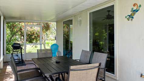 Screened patio with brick flooring, metal dining table, six mesh chairs, and a propane grill near flowering trees.