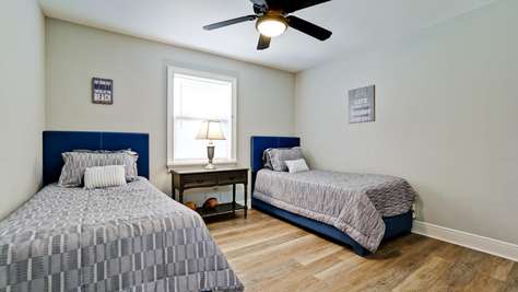 Twin beds with blue upholstered headboards flank a dark wood nightstand beneath a window.