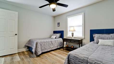 Two twin beds with blue headboards flank a dark wood nightstand under a window with white blinds.