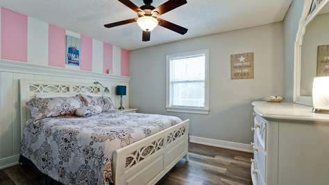 White wooden bedframe with patterned bedding sits against pink-and-white striped accent wall in bright bedroom.