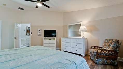 Bedroom features white dressers, a floral-patterned armchair, and a ceiling fan above wood-look flooring.