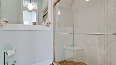 Bathroom shower with clear glass sliding doors, white square tiles, and pebble stone floor with corner shelf.