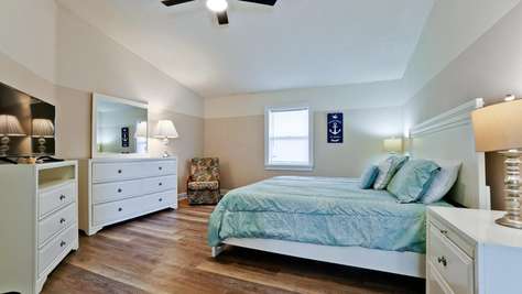 Queen bed with pale blue bedding, white wood furniture, a floral armchair, and hardwood floors.