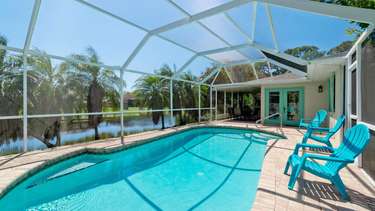 Screened pool area with turquoise Adirondack chairs on brick pavers, overlooking palm trees and a lake. - property