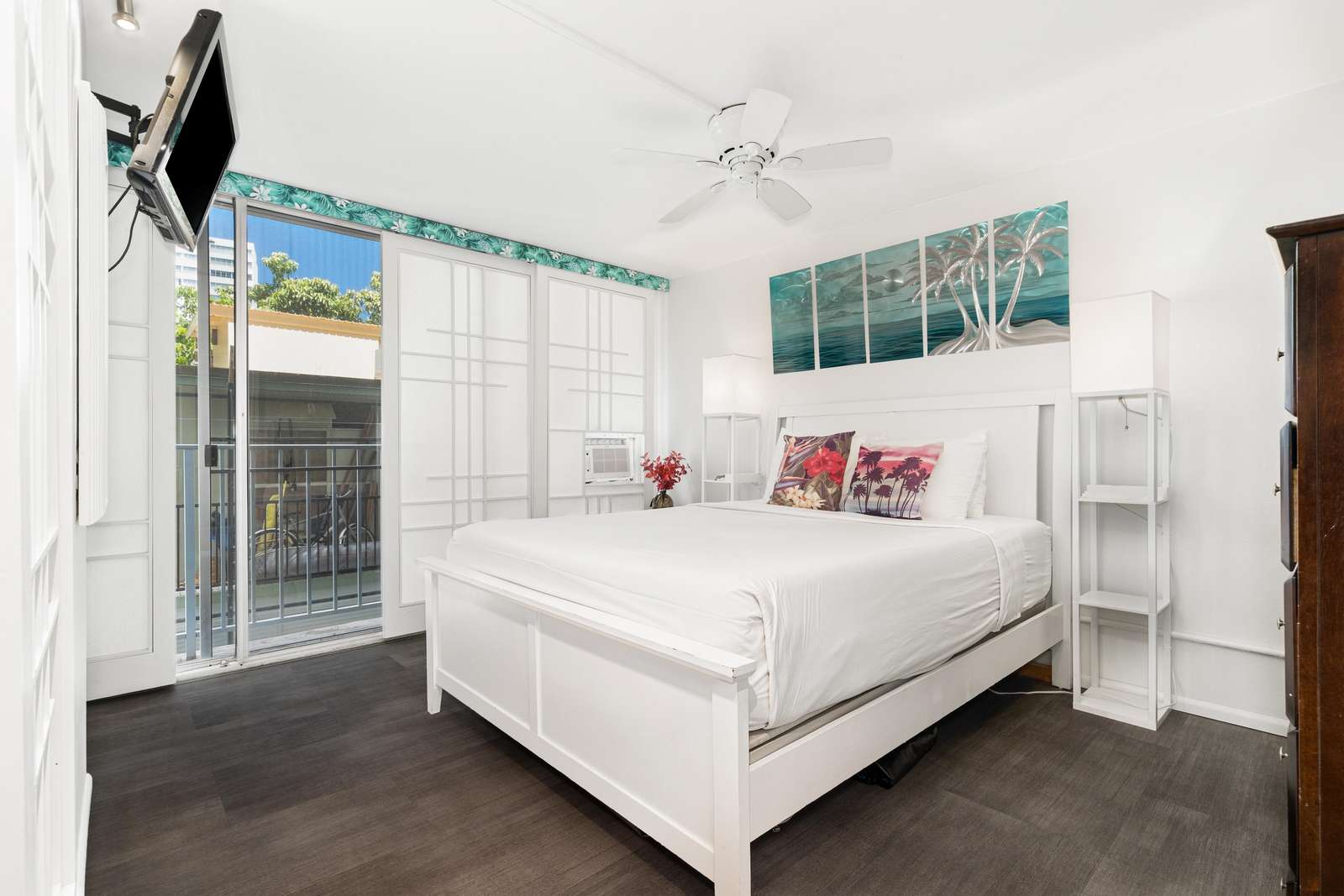 White-framed bed with tropical print pillows sits beneath a multi-panel palm tree artwork and ceiling fan.