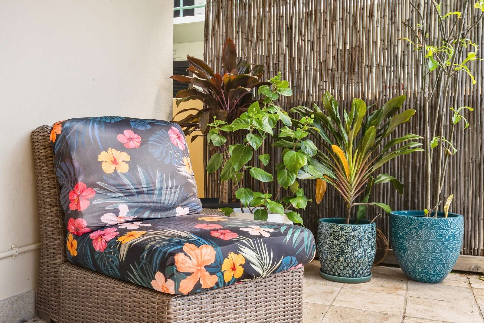 Patio corner with woven rattan chair featuring floral cushions and a bamboo privacy screen behind potted tropical plants.