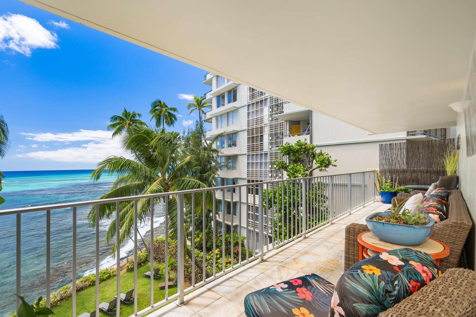 Spacious oceanfront balcony with wicker seating, floral cushions, tiled floor, and views of palm trees and turquoise water.