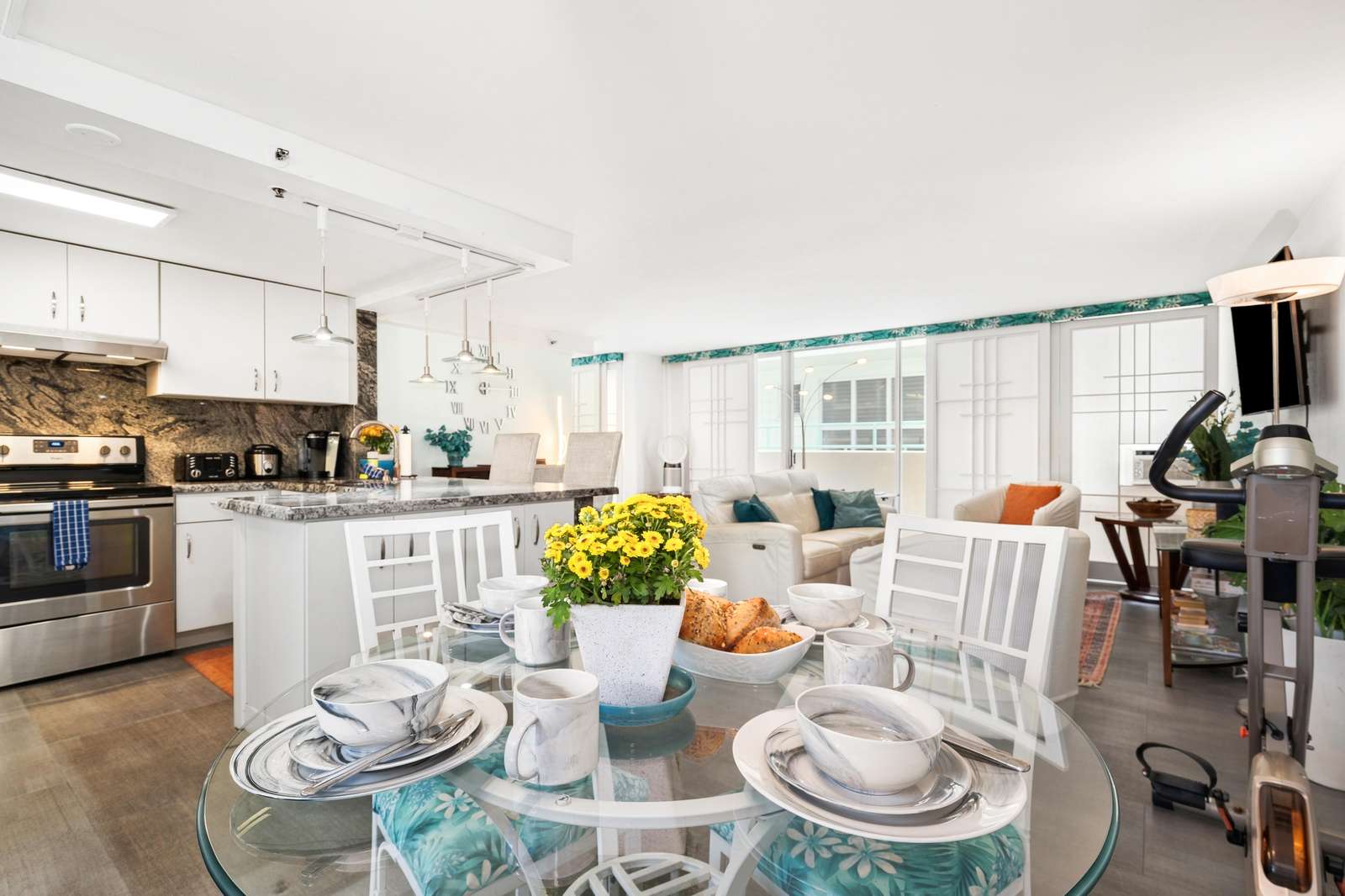 Round glass dining table with tropical cushion chairs and yellow flowers beside the white kitchen island.