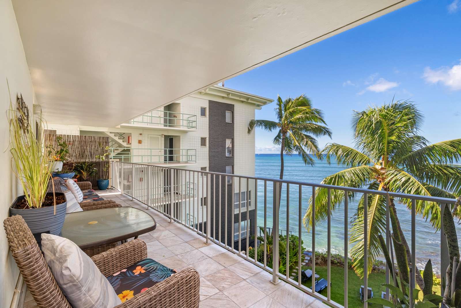 Tiled balcony with wicker chairs, floral cushions, potted plants, and partial ocean view framed by palm trees.