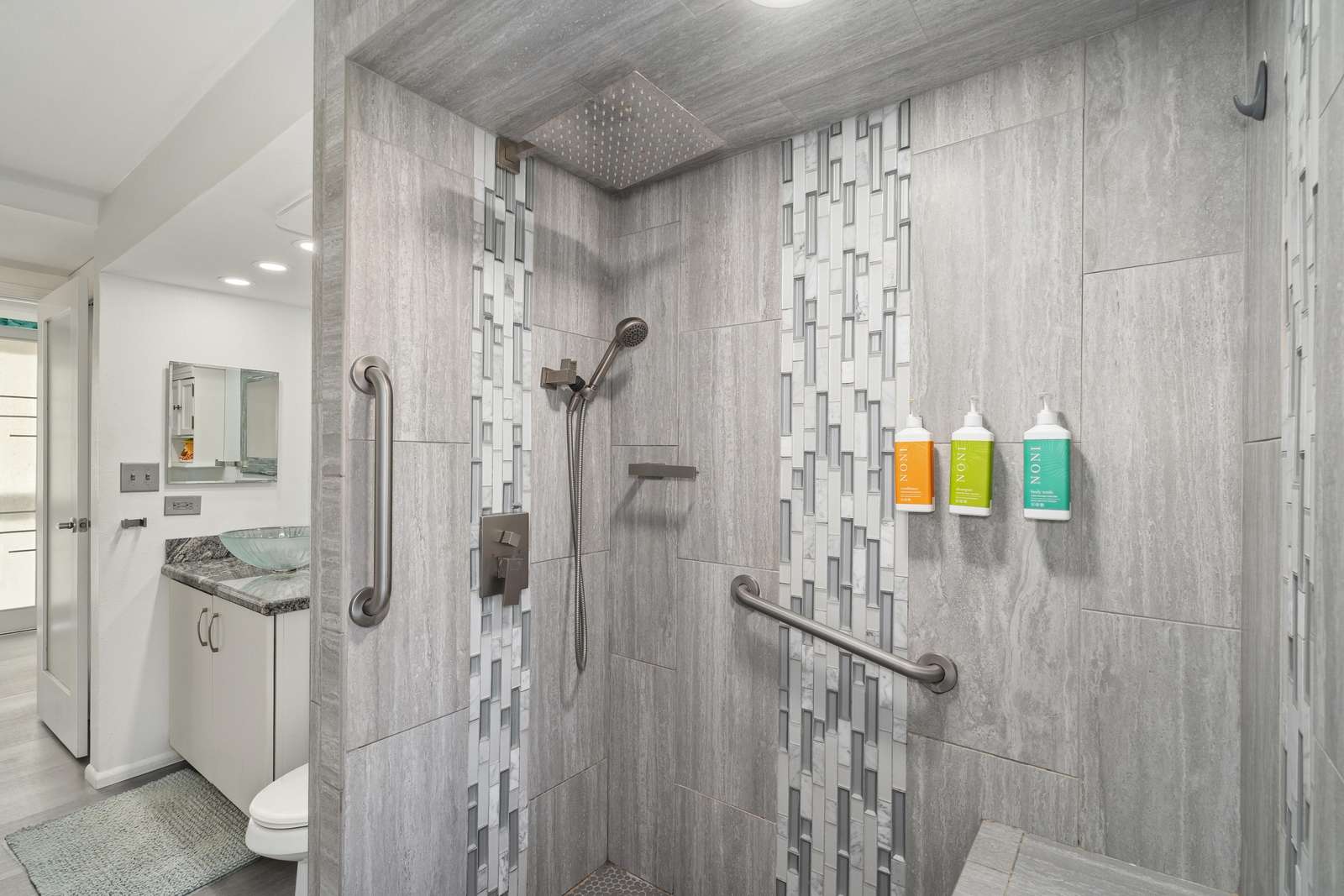 Shower with gray stone tiles, mosaic vertical accents, rain showerhead, handheld sprayer, and built-in bench.