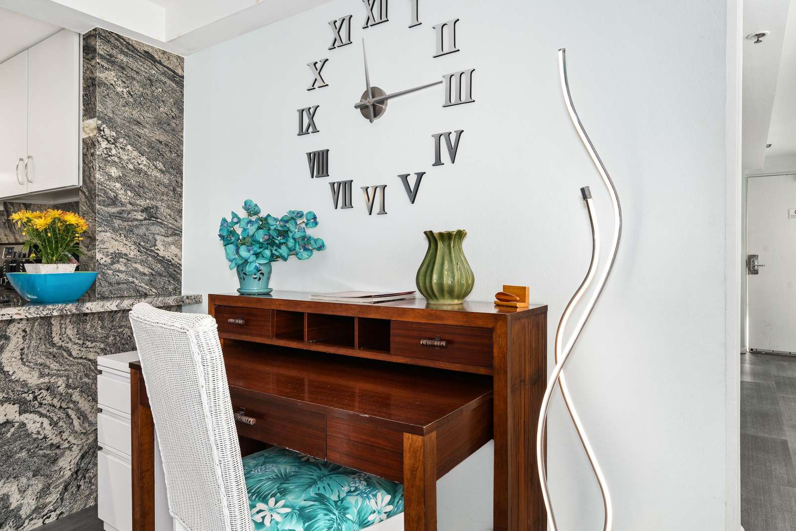 Wood desk with cubby shelves and floral-cushioned white chair beneath a wall clock with Roman numerals.