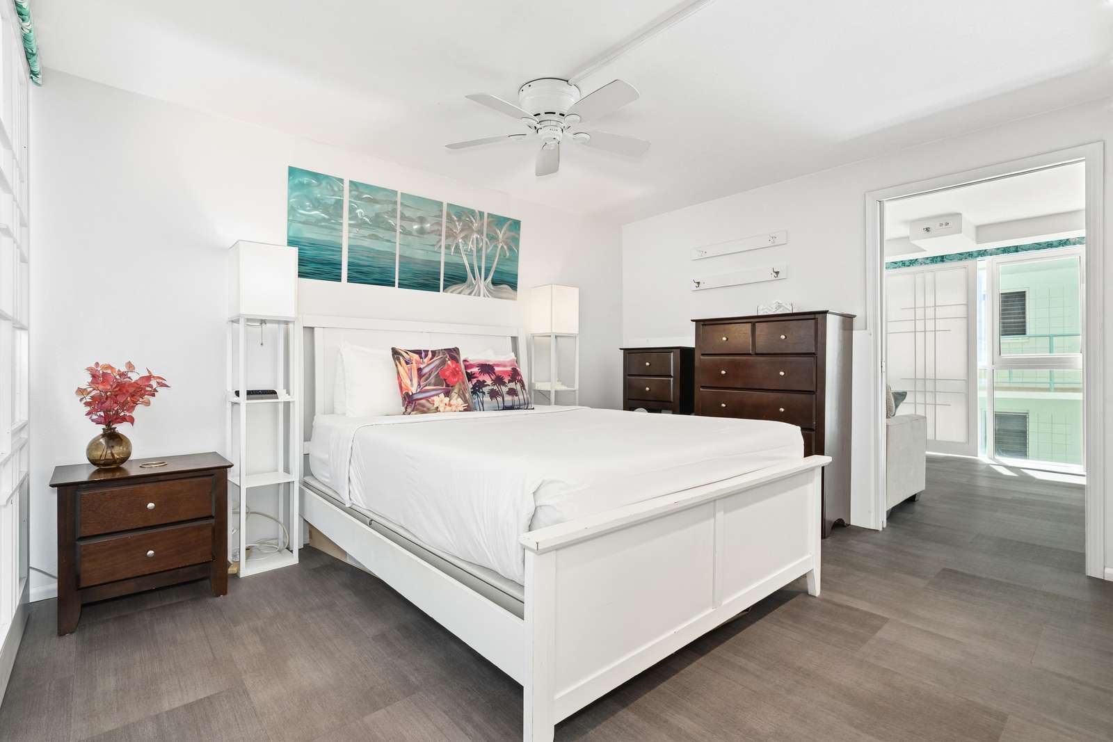 White wood bed with tropical print pillows faces dark wood dressers under a ceiling fan in the bedroom.