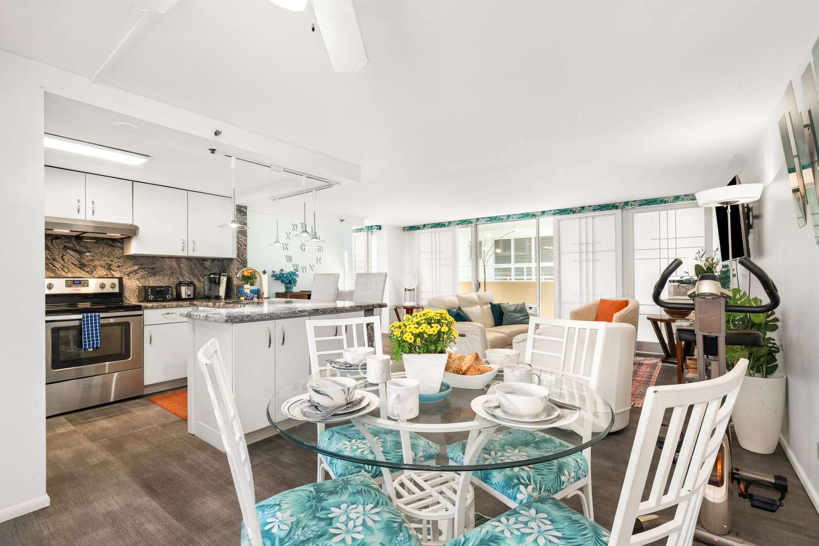 Glass-top dining table with floral cushions seats four next to a granite-topped kitchen island and white cabinetry.