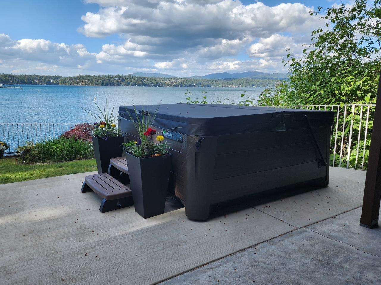 Outdoor patio features a covered black hot tub flanked by tall planters with colorful flowers and lake views.