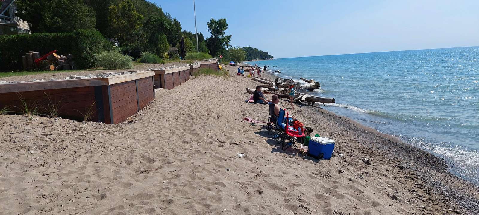 Sandy Port Albert beach lined with driftwood logs and visitors seated near calm, blue lake waters under clear skies.