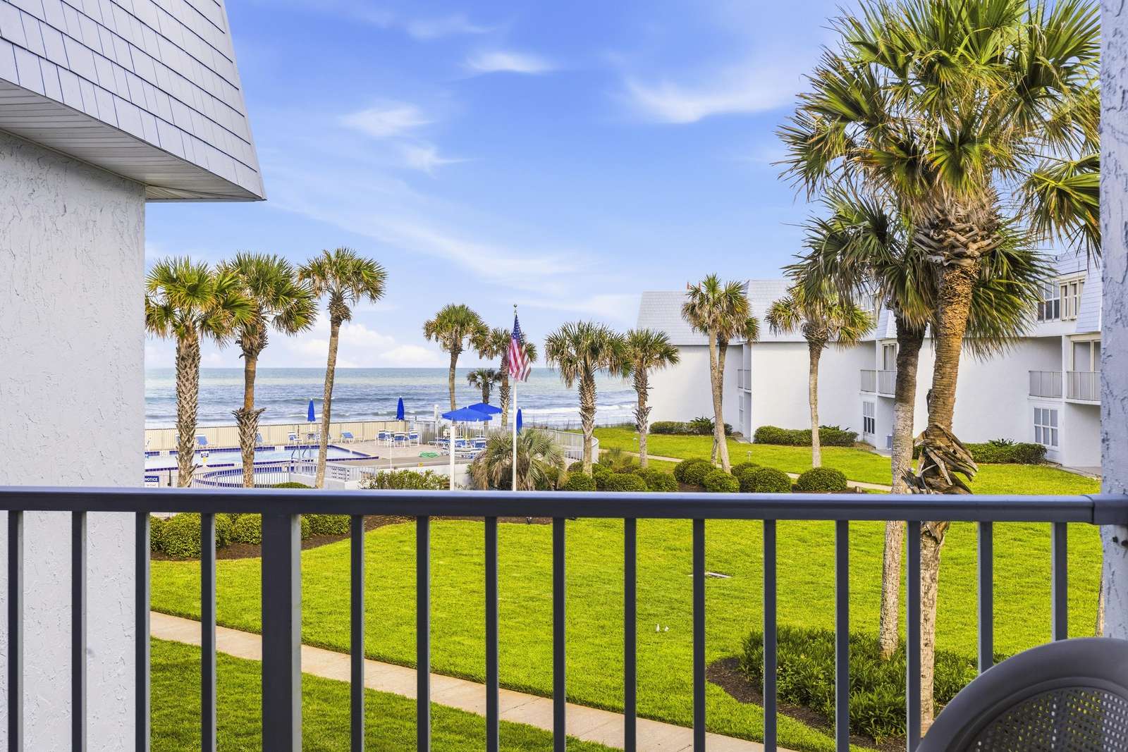 Balcony view overlooks manicured lawn, tall palm trees, a fenced pool area with blue umbrellas, and ocean beyond. - property