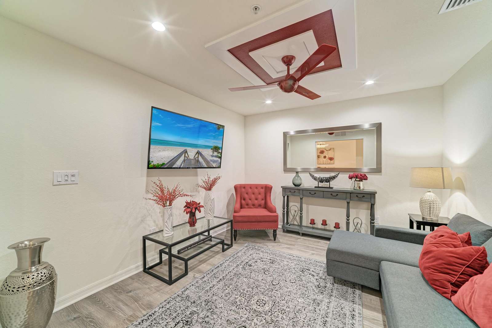 Living area with gray sectional sofa, red accent pillows, tufted armchair, glass coffee table, and wall-mounted TV. - property