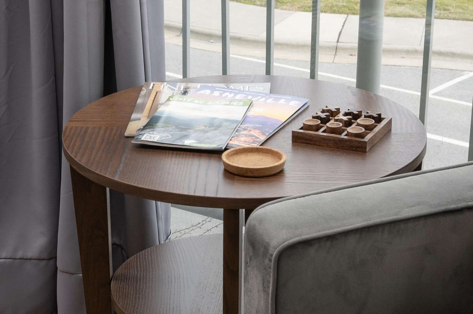 A round wooden table with magazines and a game on it, next to a gray chair in front of a window.