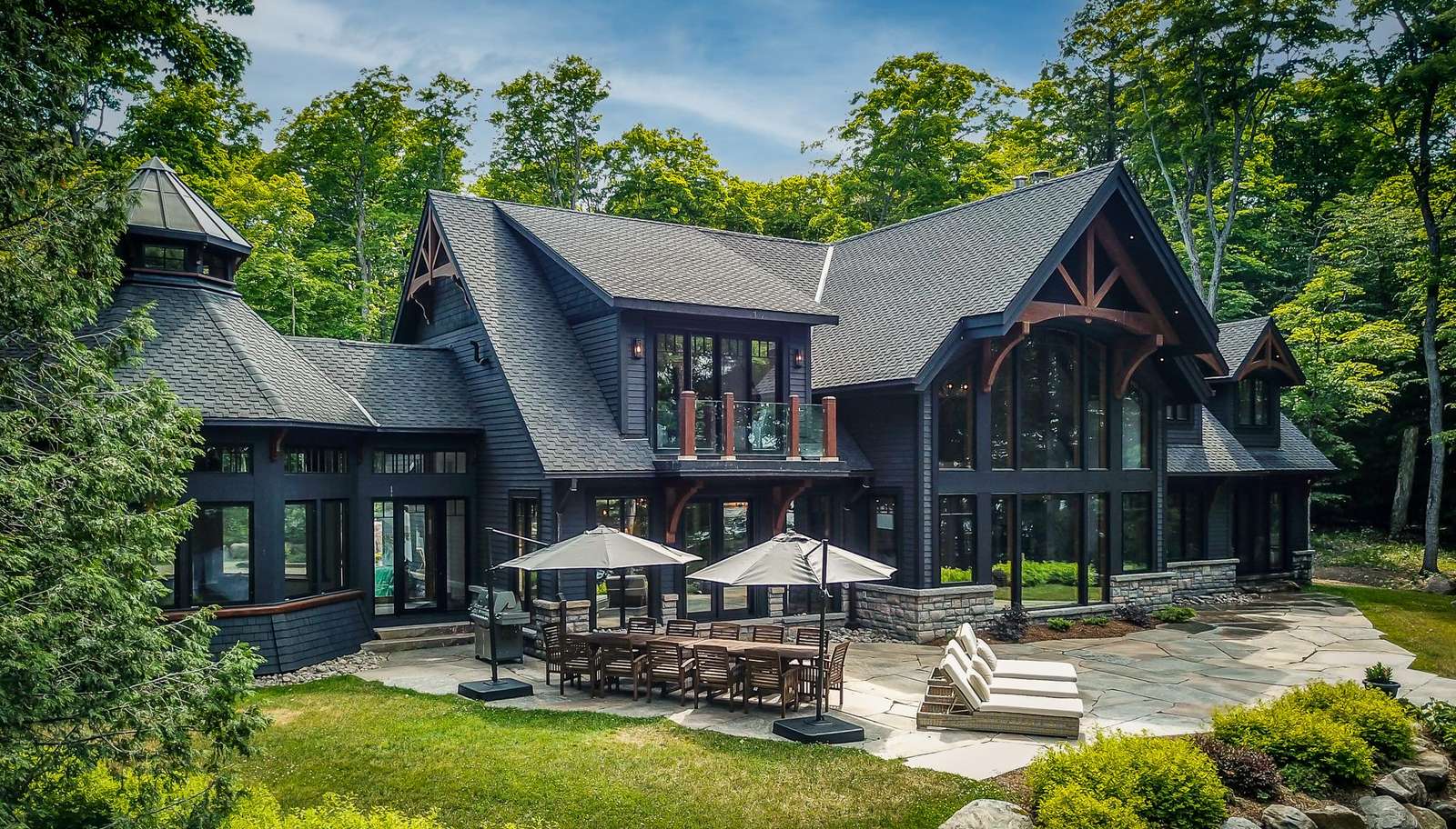 Stone patio with wooden dining set and umbrellas, flanked by cushioned lounge chairs beside dark wood cottage. - property