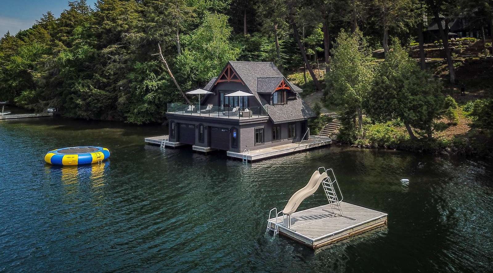 Dark wood boathouse with rooftop deck, glass railing, and lakeside dock featuring a floating slide and trampoline.