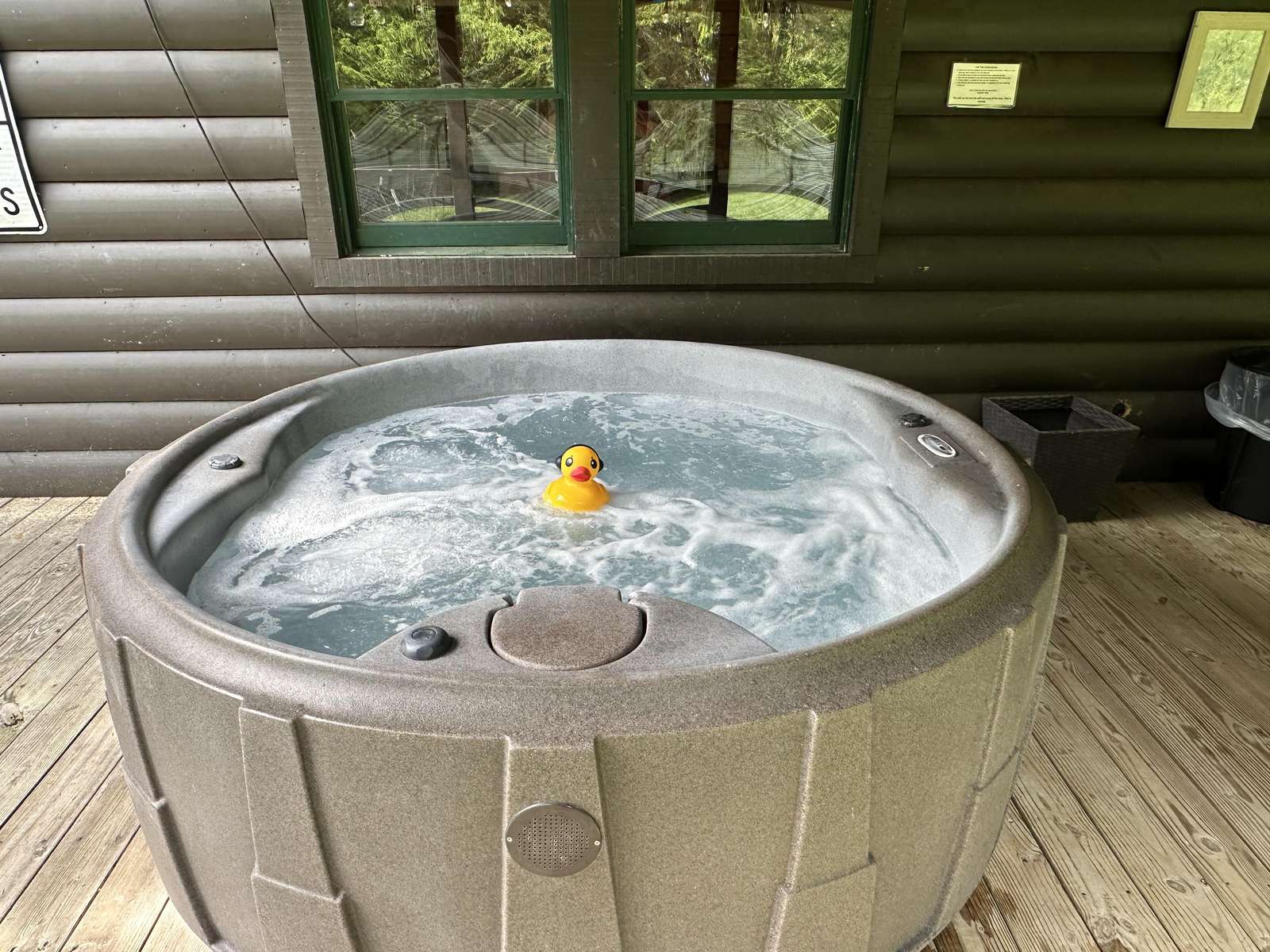 Outdoor hot tub bubbling with a yellow rubber duck, set on a wooden deck against dark log cabin walls. - property
