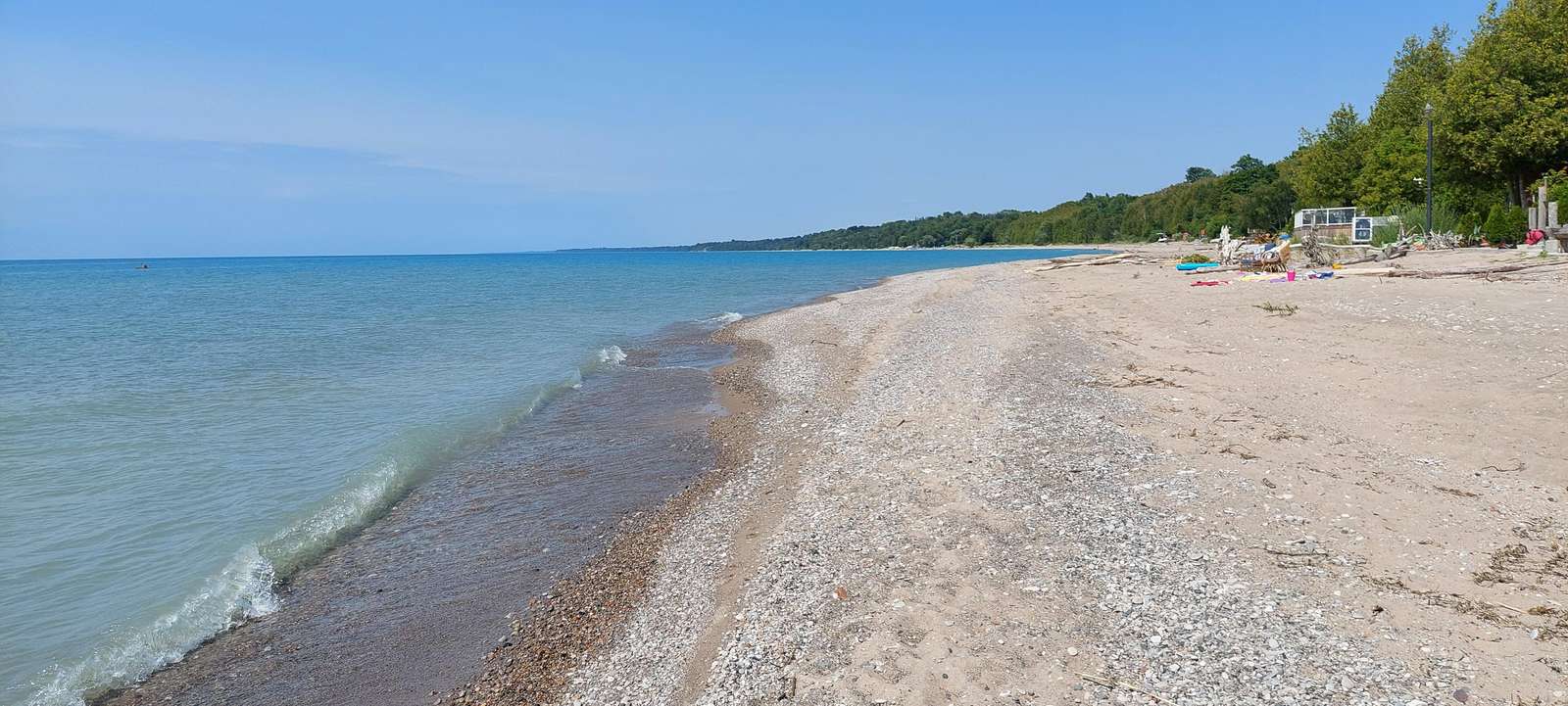 Sandy and pebbled shoreline curves alongside calm blue waters under a clear sky with trees lining the distant shore.