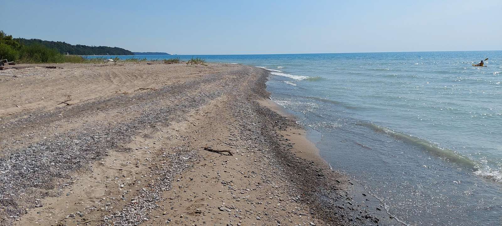 Sandy and pebbled shoreline curves alongside calm blue waters with a kayaker paddling near the horizon.