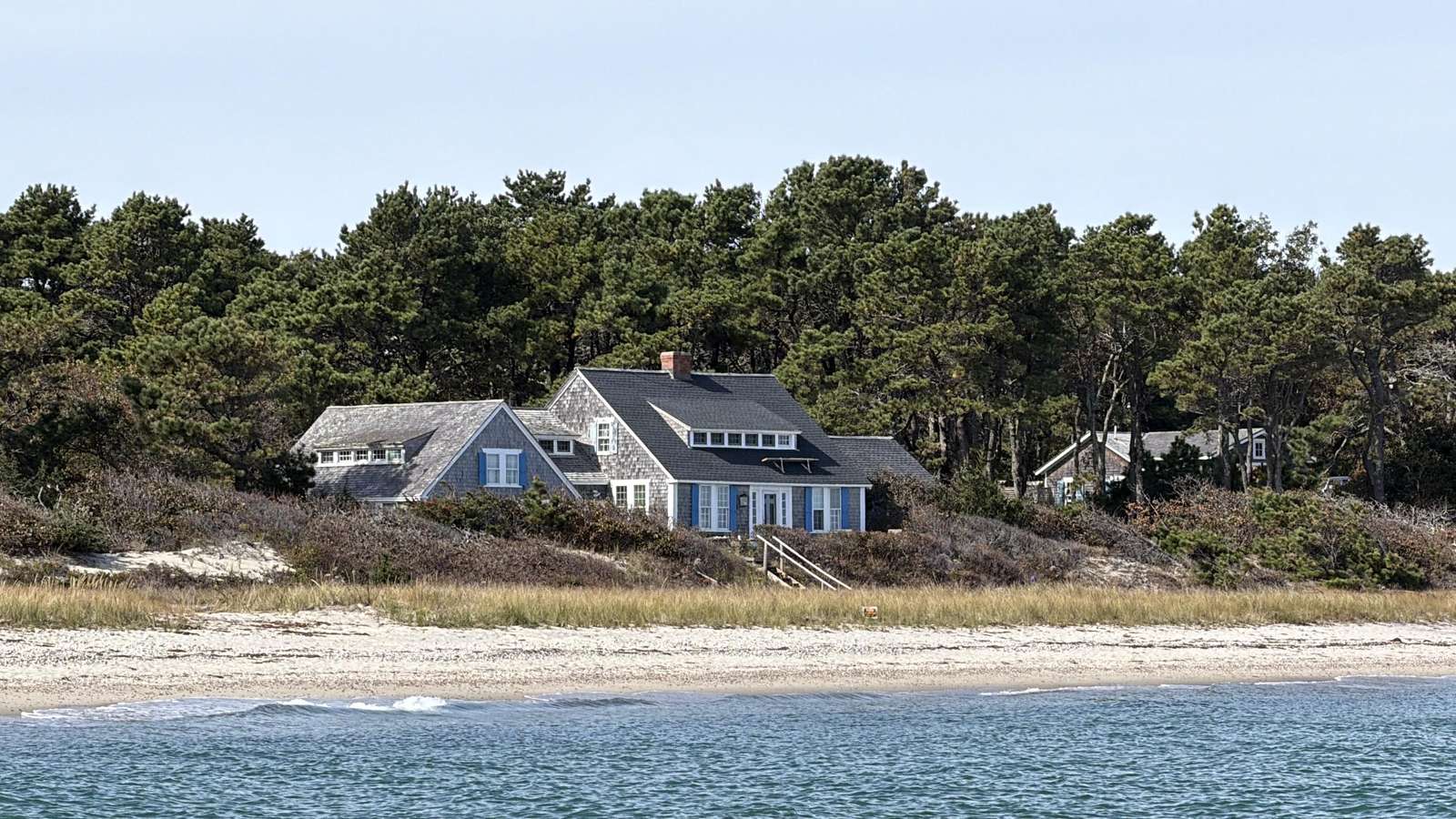 Shingled beachfront house with blue shutters and multiple gables nestled behind dune grass and pine trees. - property