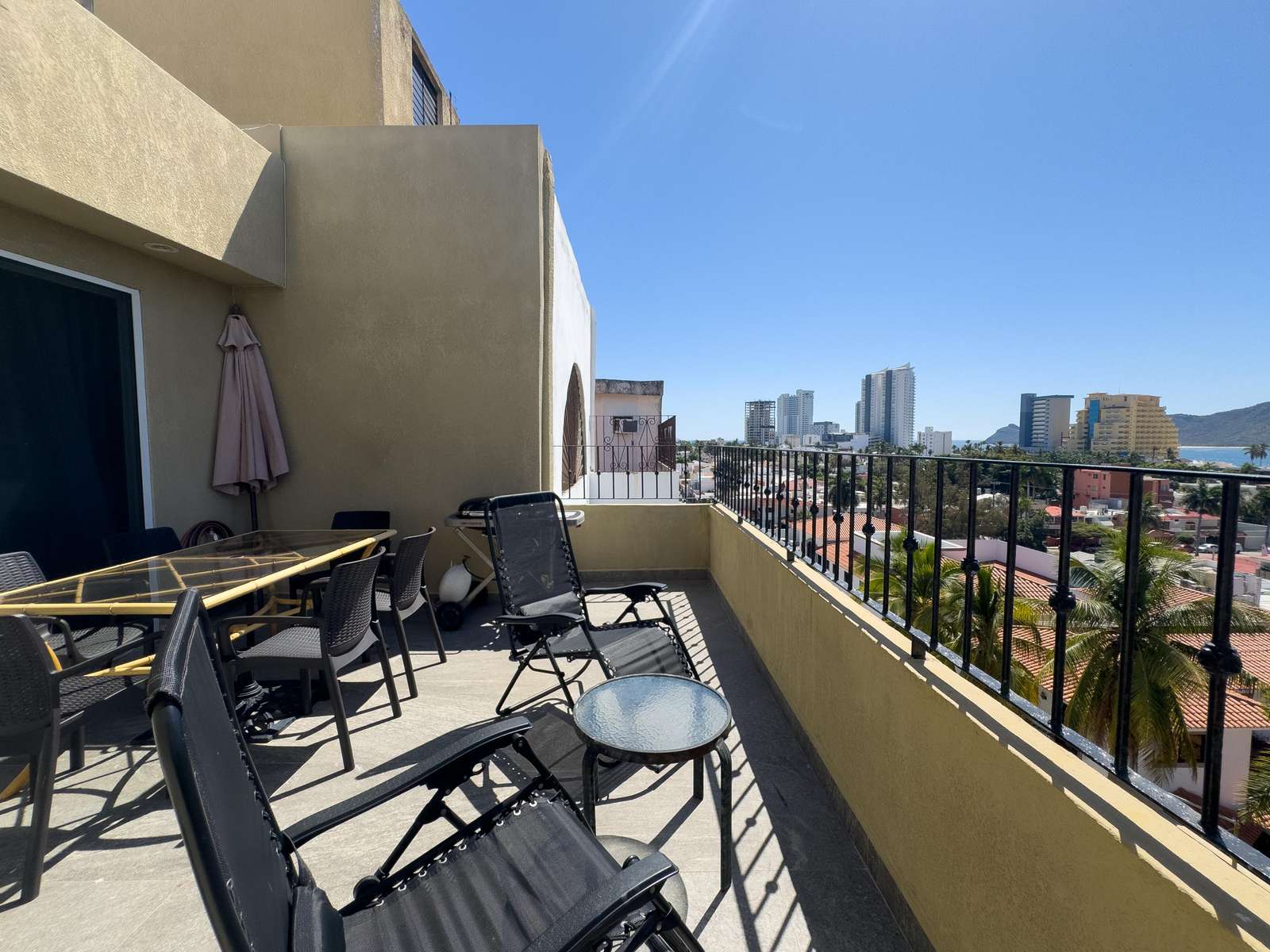Spacious balcony with black metal railing, glass-top dining table, lounge chairs, and city skyline views. - property