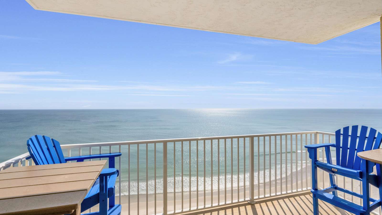 Balcony with blue Adirondack chairs and tan wood tables overlooks sandy beach and shimmering ocean. - property