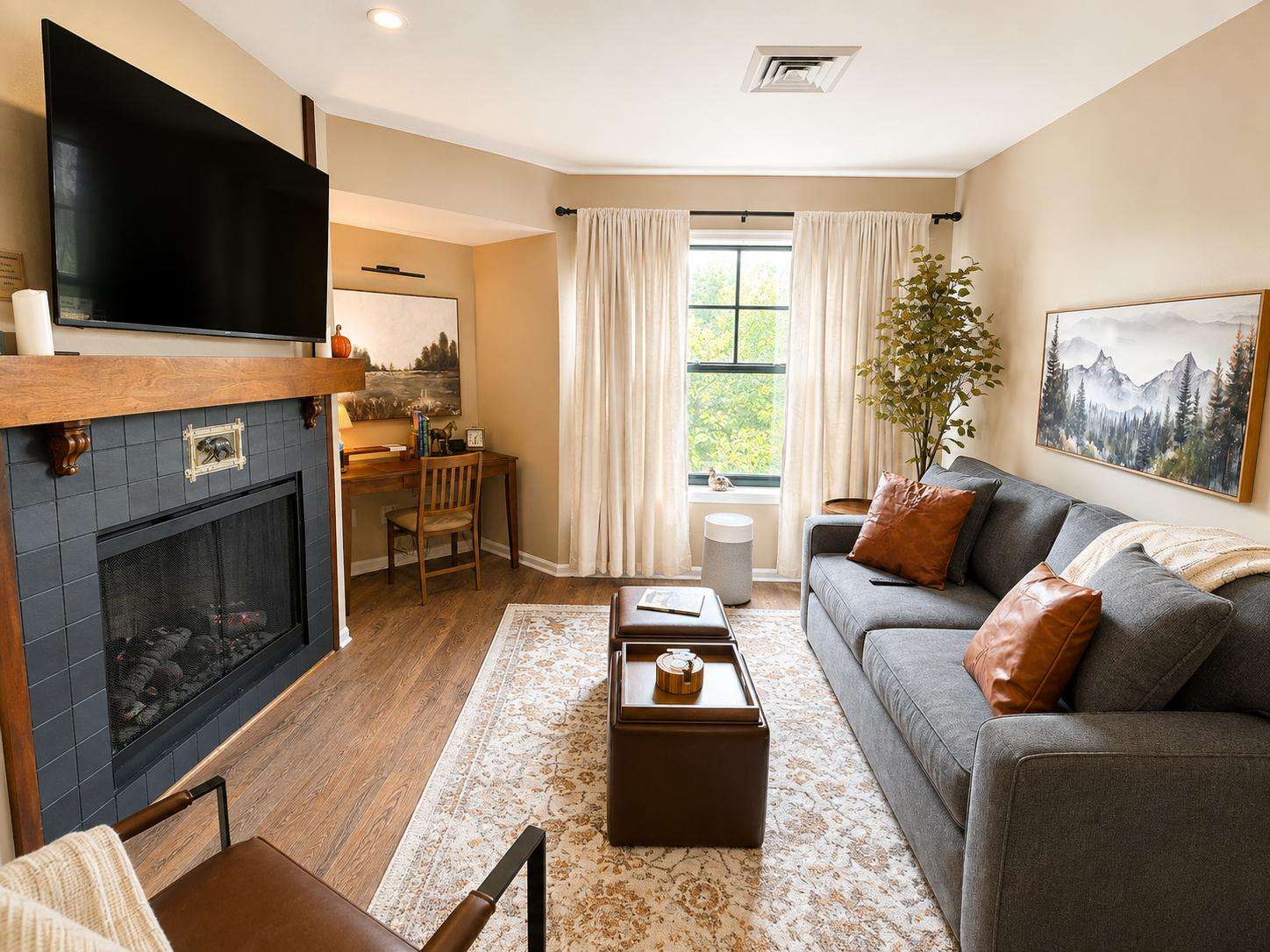 Living area with gray sofa, leather pillows, wooden desk, black-tiled fireplace, and window framed by cream curtains. - property