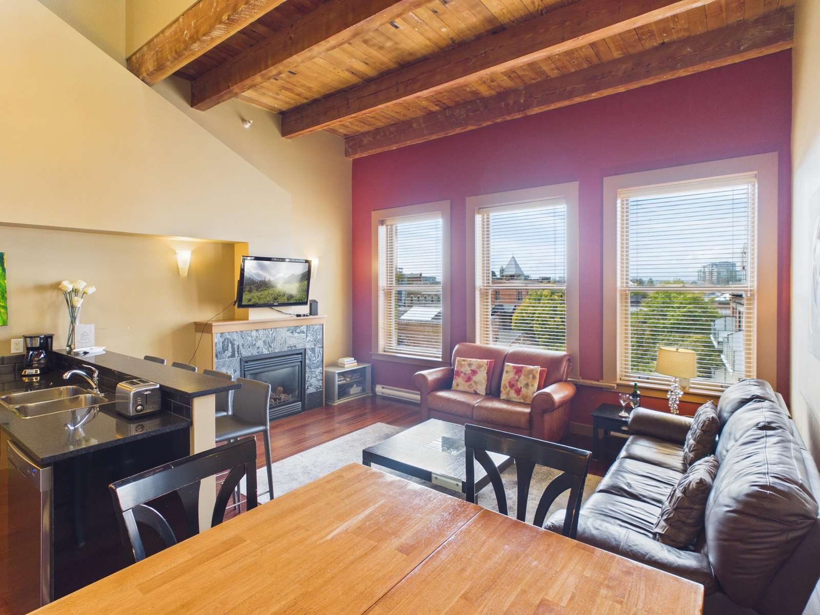 Living room with exposed wood beams, three tall windows, leather sofas, stone-tiled fireplace, and black kitchen counter. - property